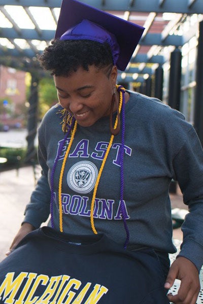 Glenesha Berryman, wearing graduation hat & honor cords and an East Carolina University sweatshirt while admiring a Michigan University sweatshirt outside of the Wright Building.